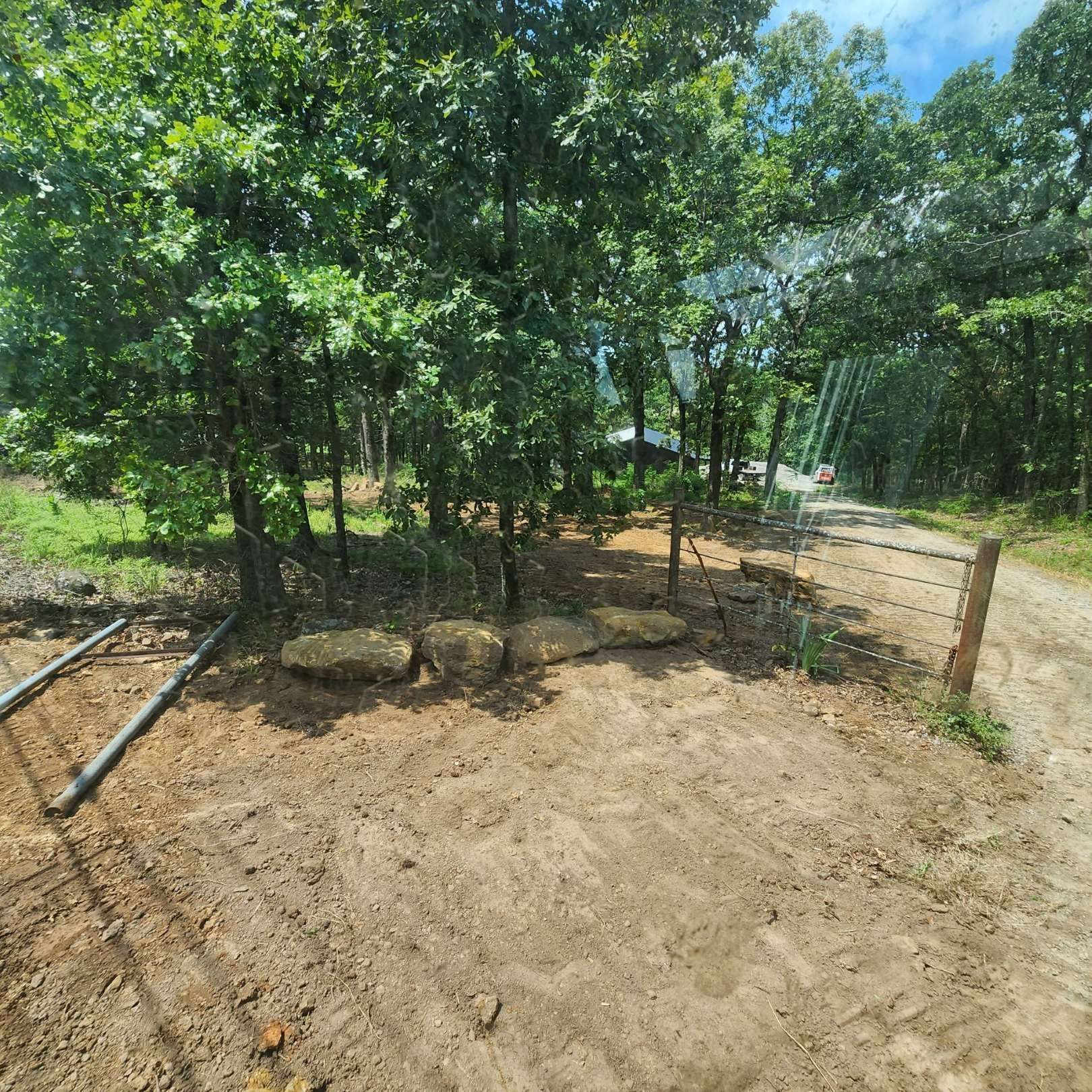 Dirt driveway entrance lined with large rocks and trees; fence on the right.