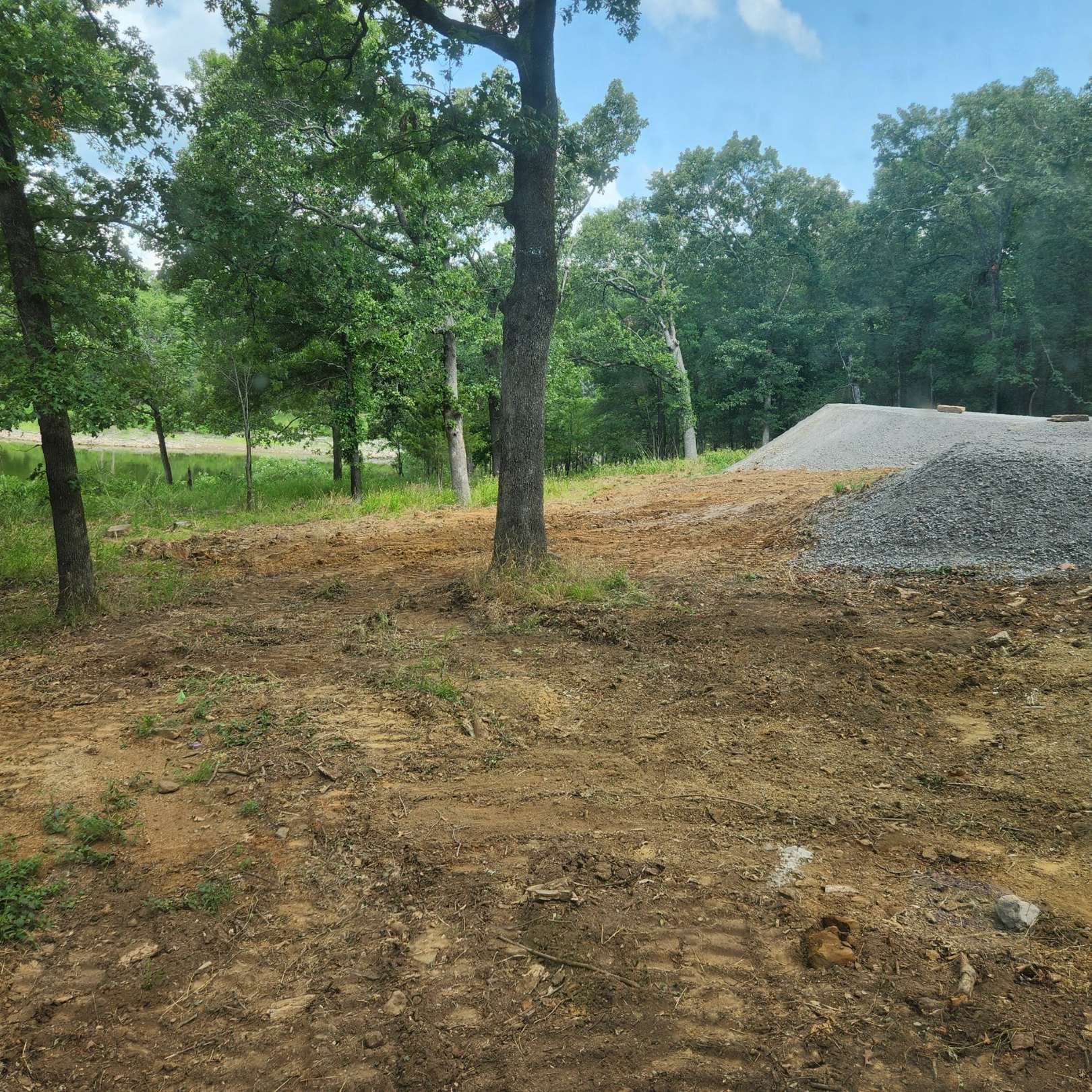 Dirt clearing with trees and a pile of gravel in a wooded area under a cloudy sky.