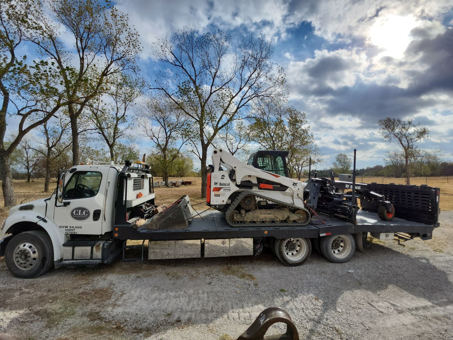 A white flatbed truck with a skid steer loader on the bed, outdoors on a cloudy day.