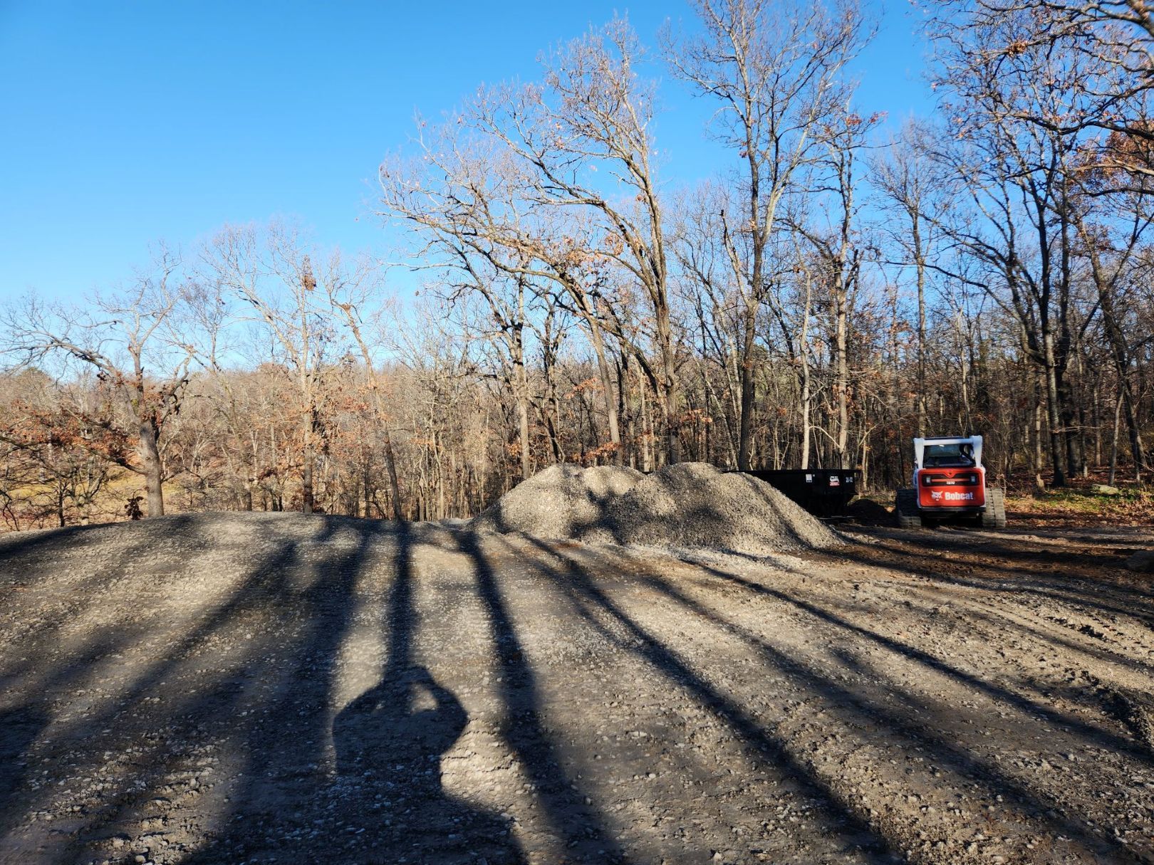 Shadowed gravel surface with trees, a small pile of gravel, and a red skid steer on a sunny day.