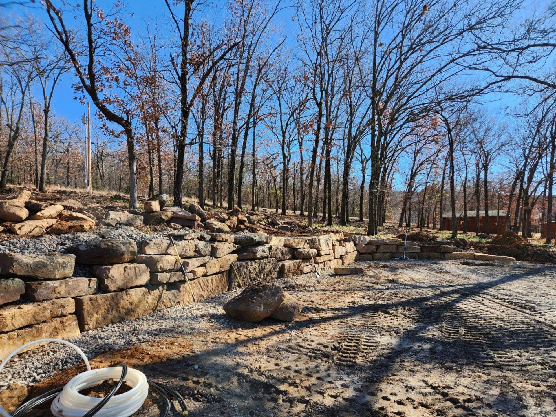Stone retaining walls and gravel driveway in a wooded area under a blue sky. Bare trees.