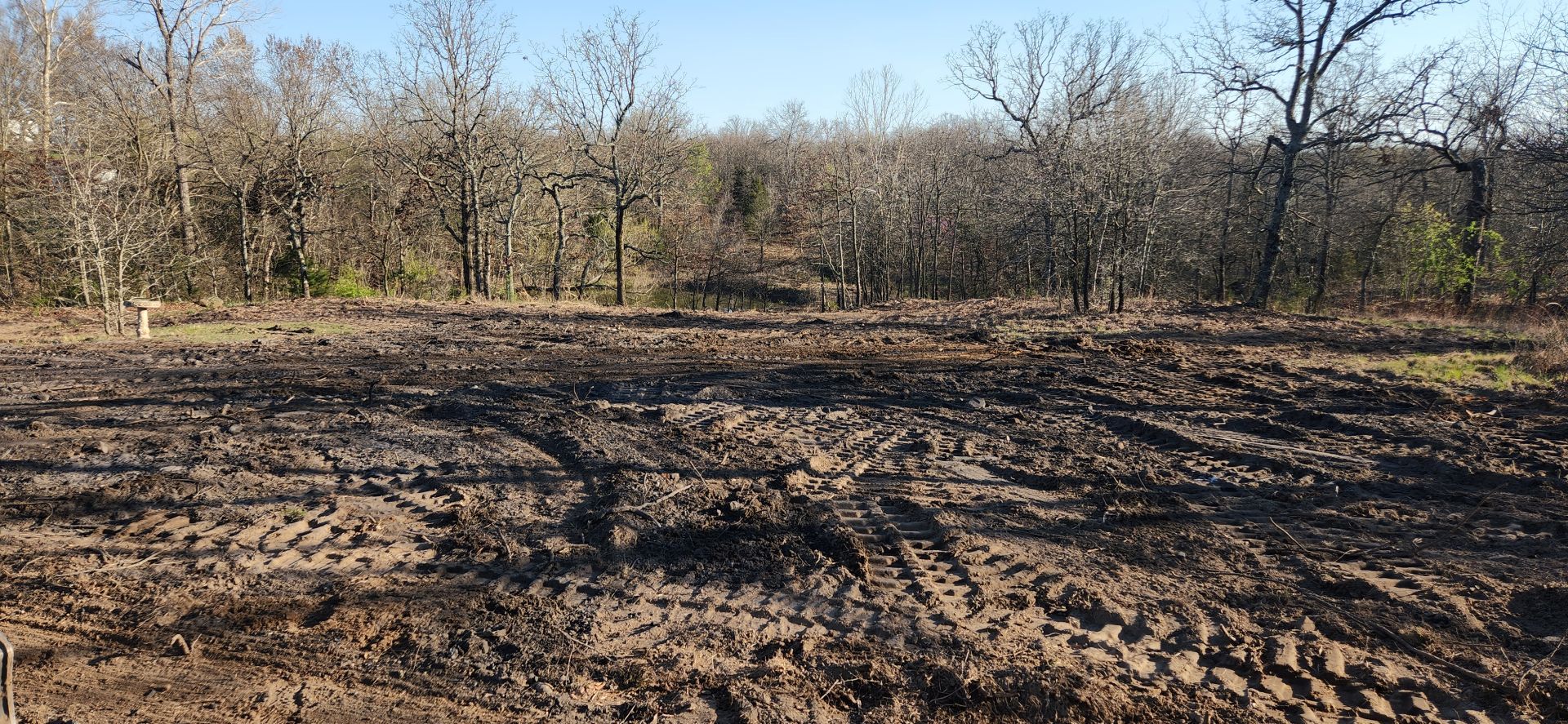 Dirt field with tire tracks and trees in the background under a blue sky.