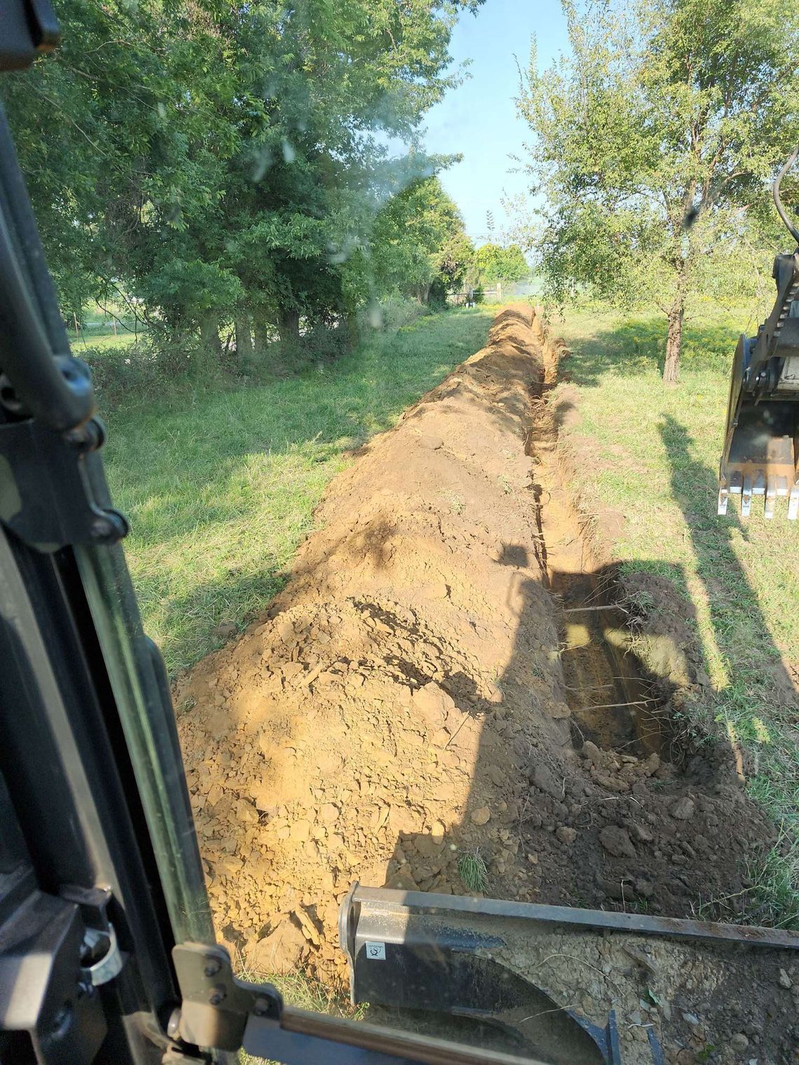 A trench in a grassy area, likely for utility lines, seen from the cab of an excavator.