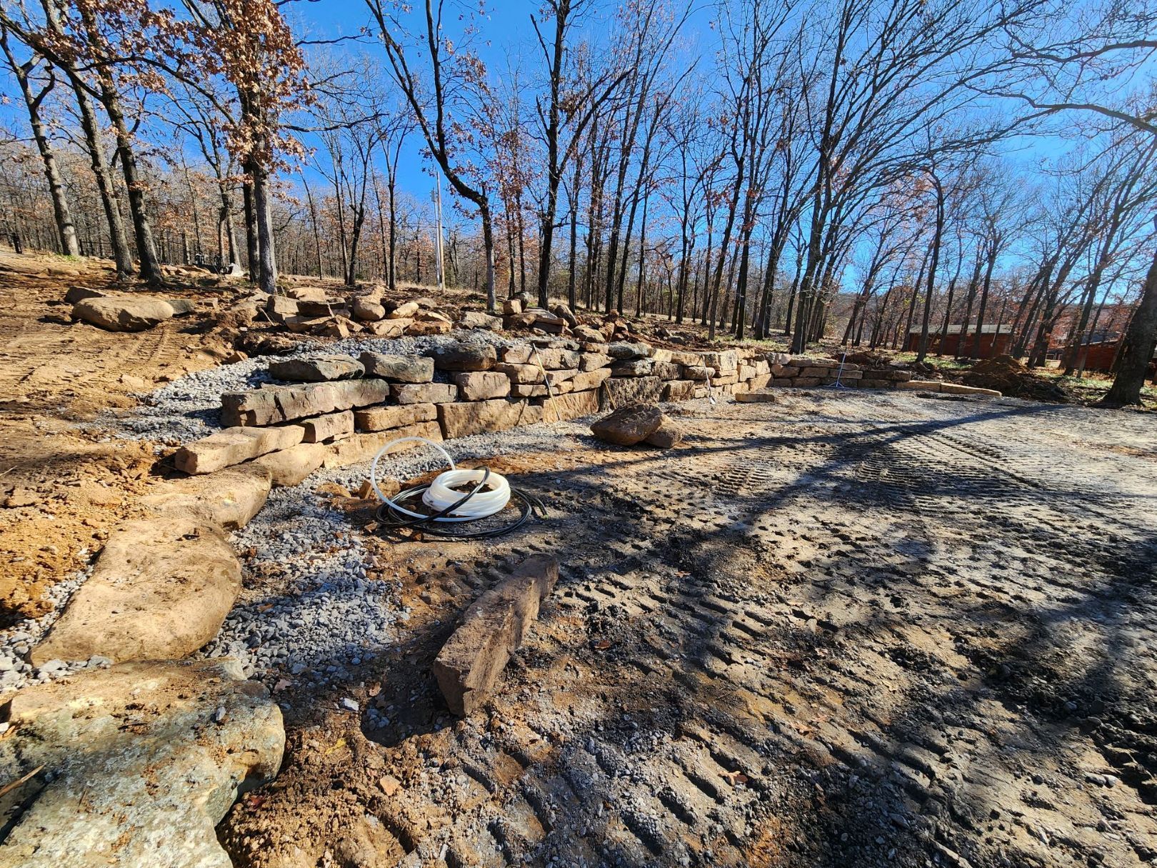 Stone retaining wall being built in a wooded area, with dirt and exposed roots.
