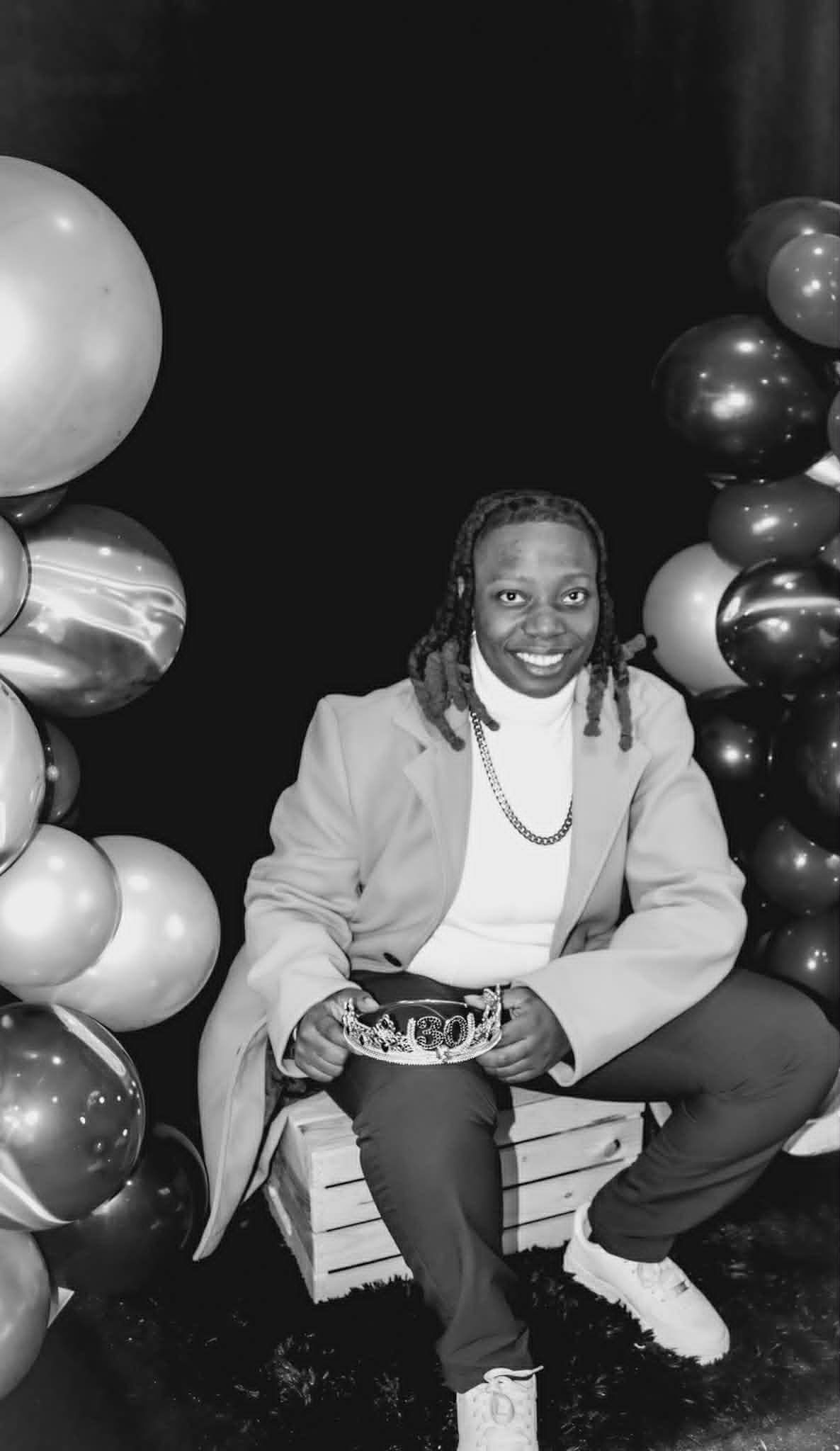 Person seated on a crate, smiling beside balloon clusters in a dark party setting