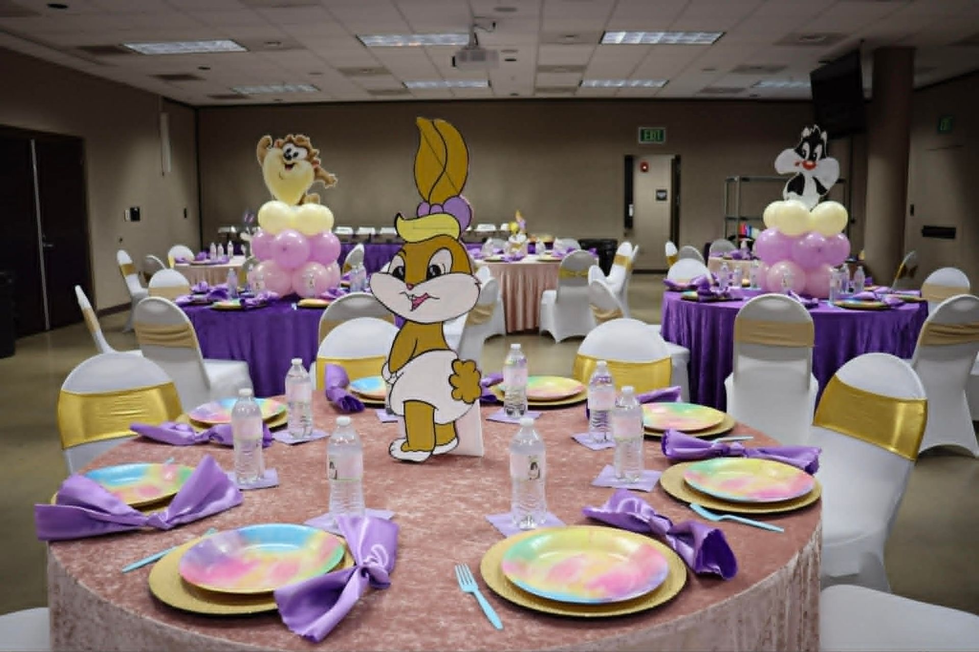 Party table with purple decorations, pastel plates, and a cartoon cutout centerpiece in a banquet room