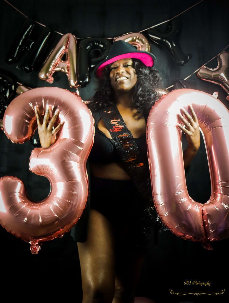Woman posing with pink “30” balloons and a matching pink hat at a birthday celebration