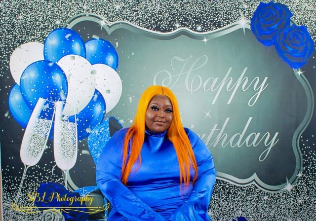 Woman in blue dress with orange hair posing before a silver “Happy Birthday” backdrop and blue-white balloons