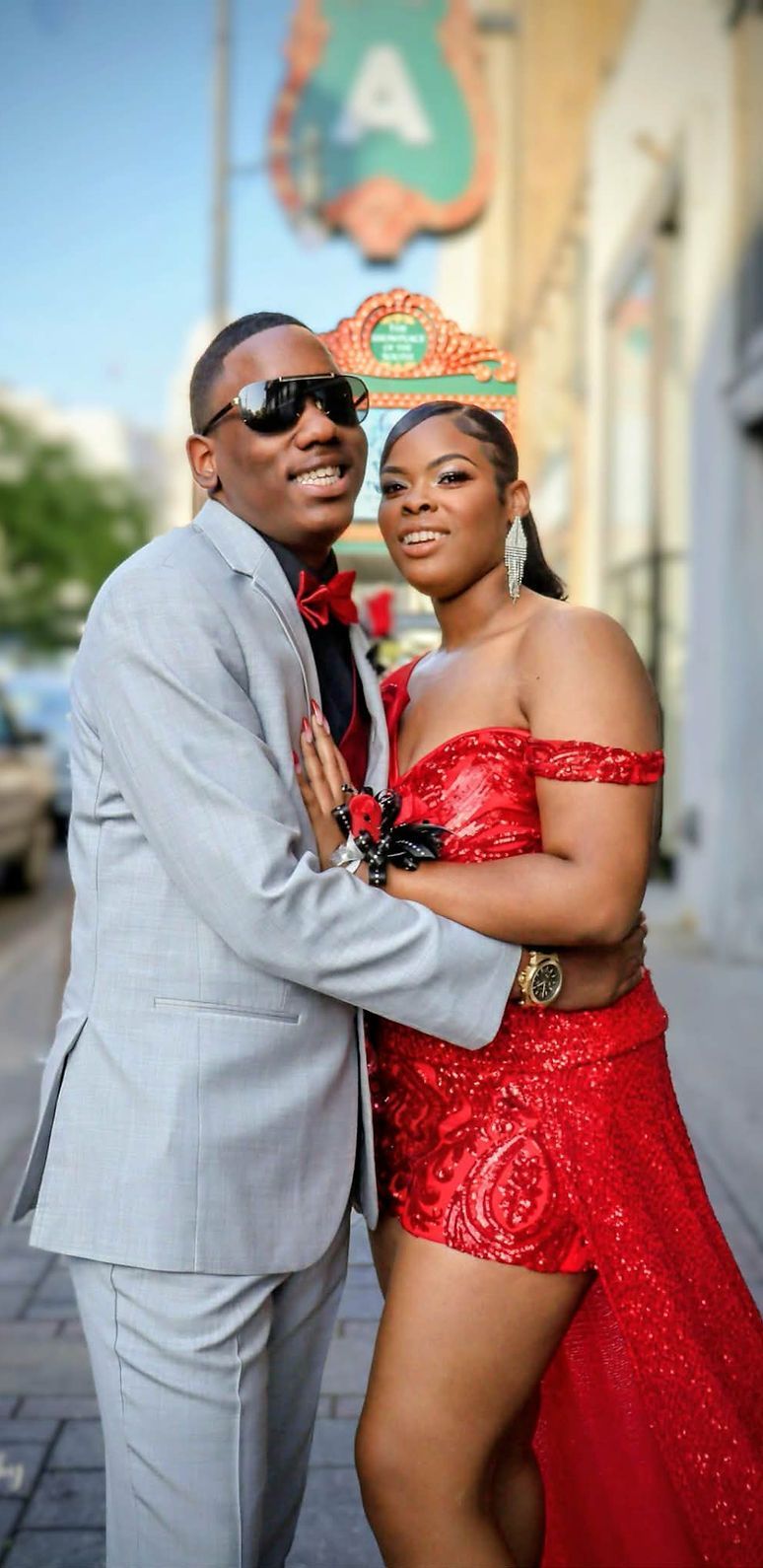 Couple posing outdoors, smiling in coordinated formal attire, with a blurred city street behind them