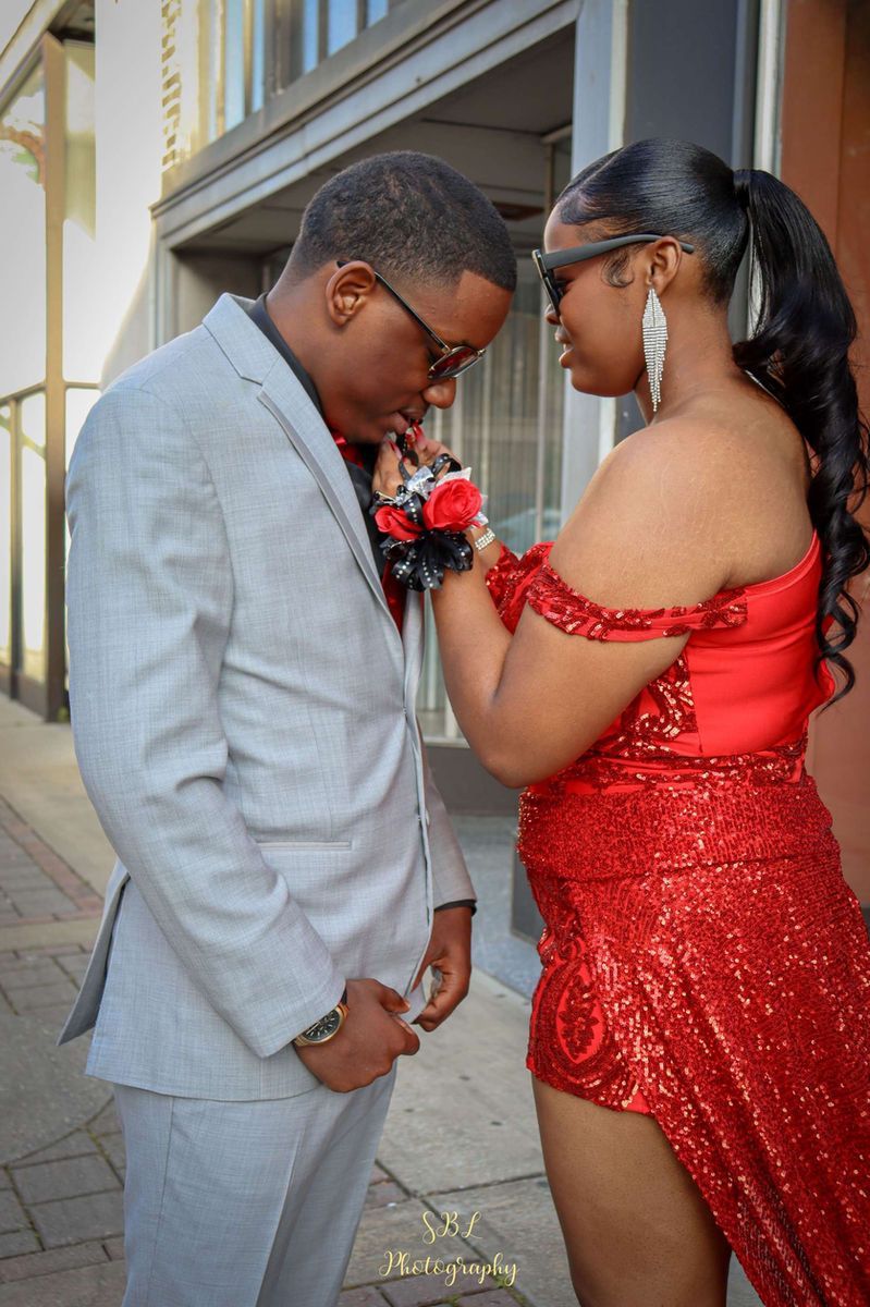 Two people in formal attire face each other, holding hands, outdoors by a building.