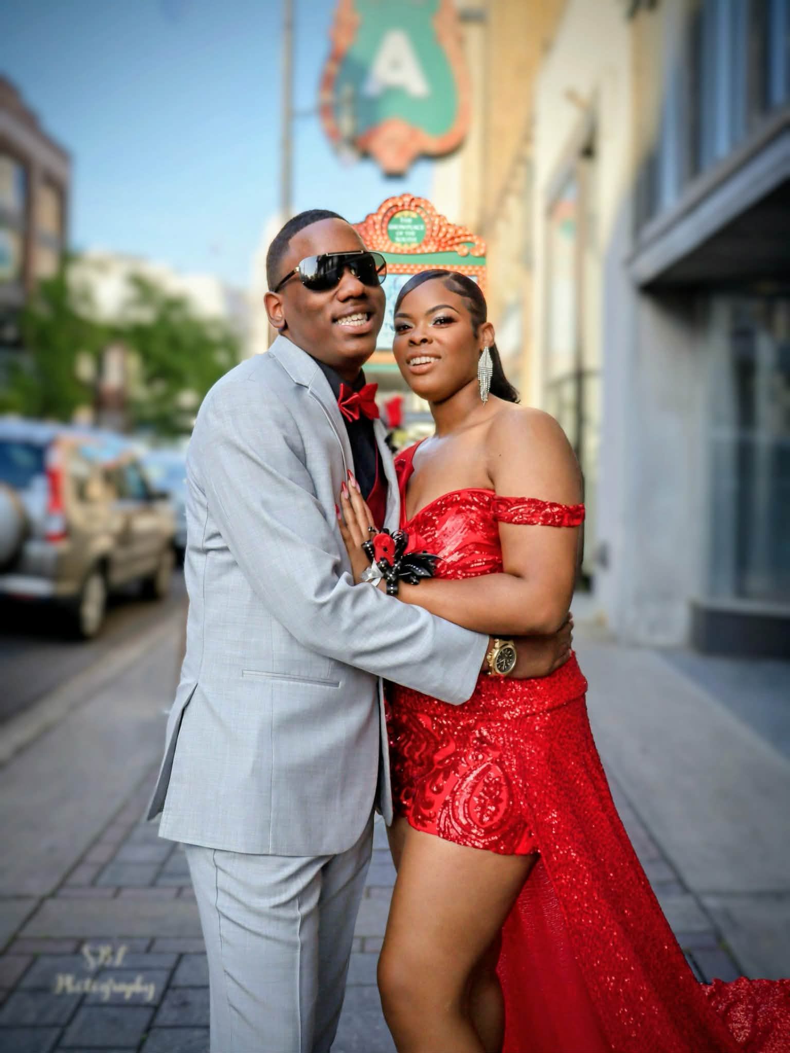A couple in formal attire posing on a city street, the woman in a red sequined dress and the man in a light gray suit.