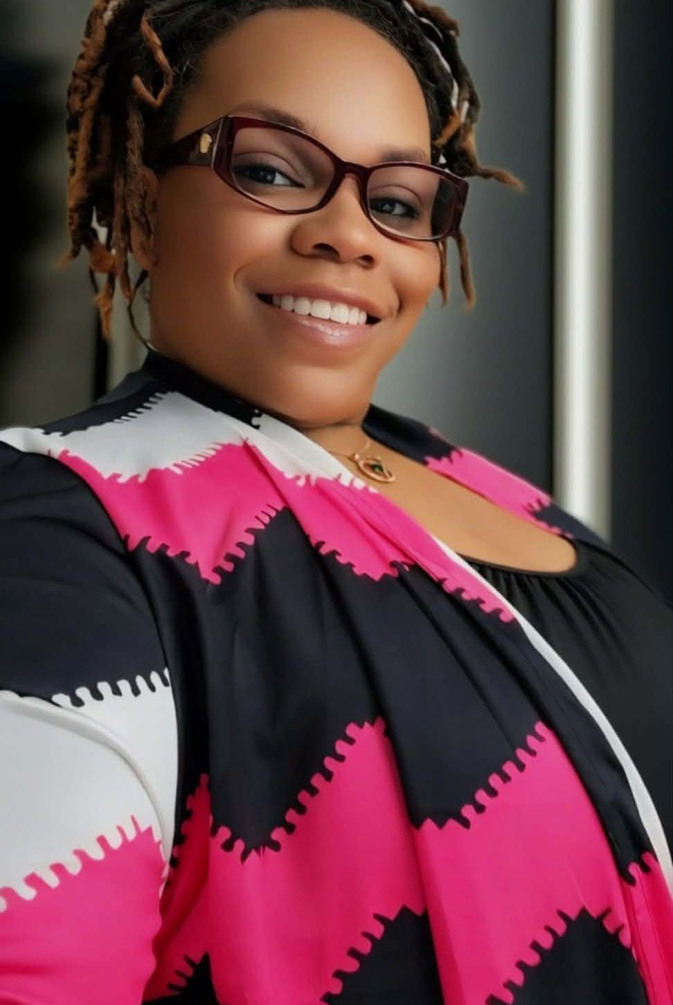 Smiling person in glasses wearing a black, pink, and white patterned jacket