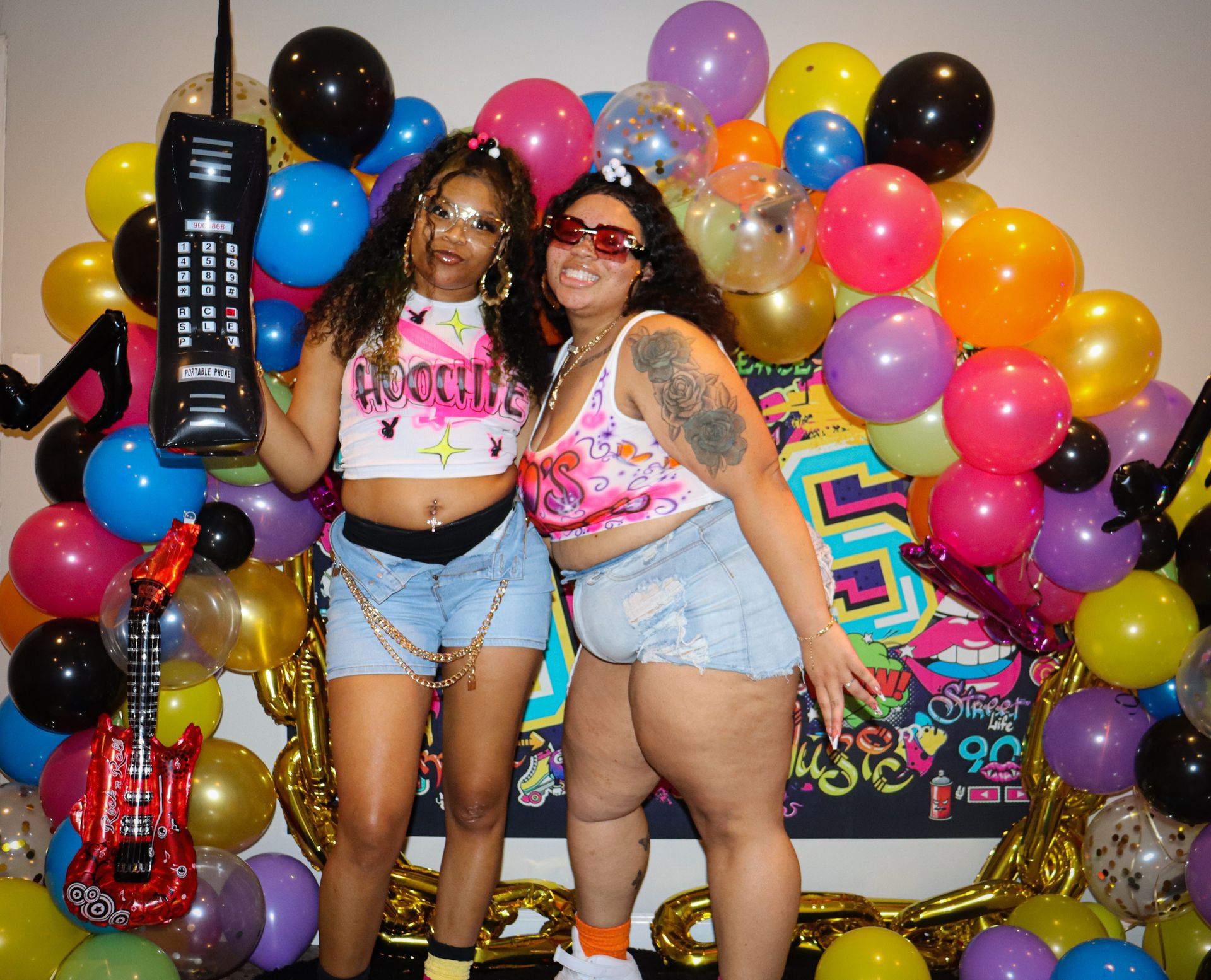 Two women posing in front of colorful balloons at a party, smiling and wearing matching pink tops and denim shorts