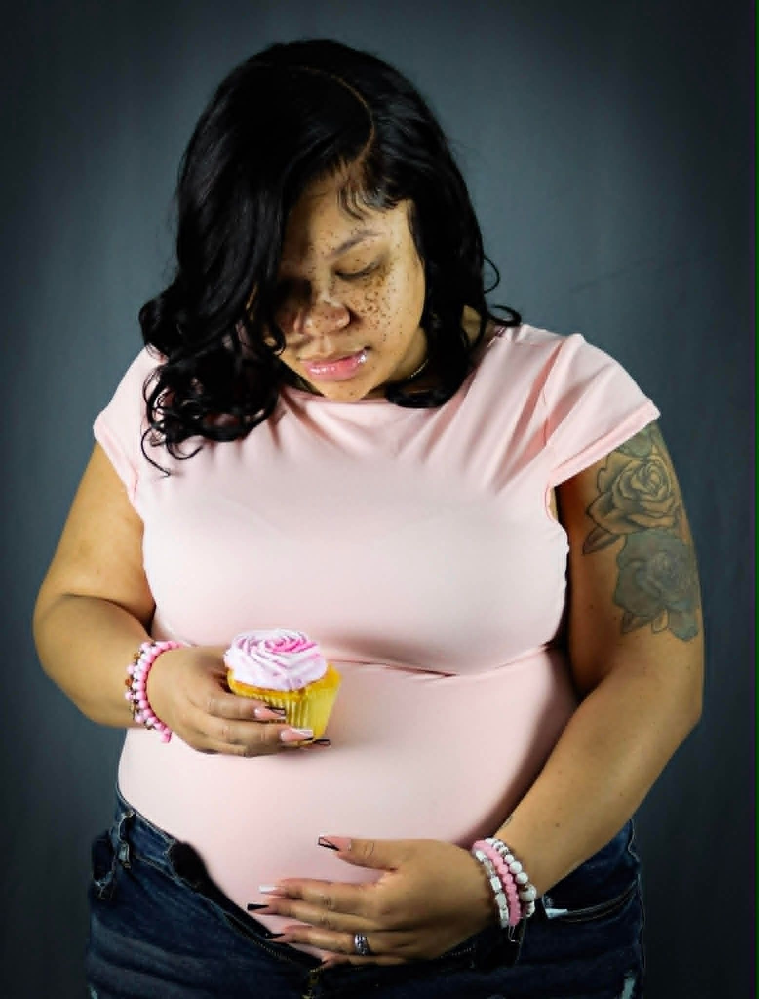 Woman in a pink shirt holding a cupcake and looking down at her pregnant belly