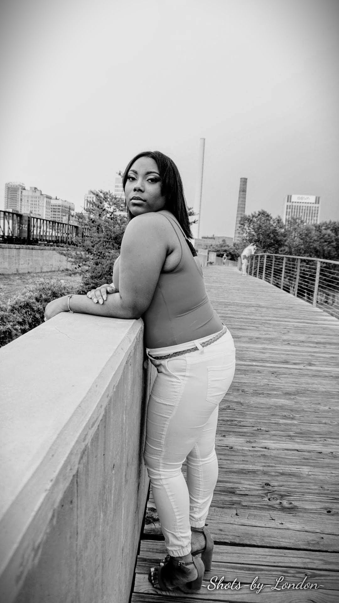 Person leaning on a bridge railing, looking over a wooden boardwalk in black and white.