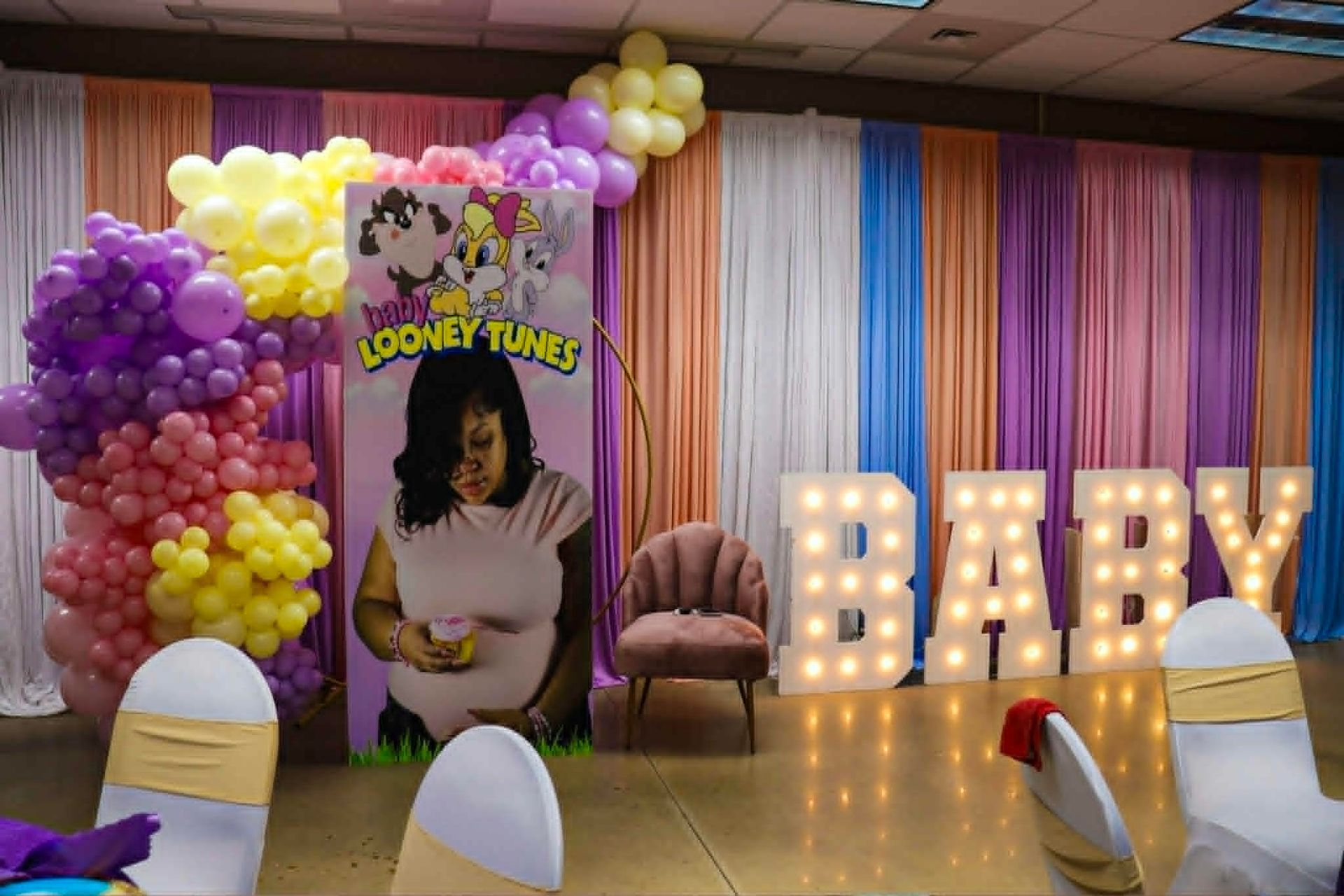 Baby shower backdrop with purple, pink, and white balloons, lit “BABY” letters, and a chair.