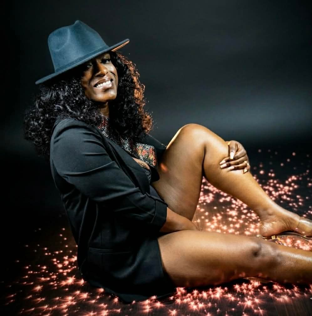 Smiling person in a black suit and hat sitting on a glowing floor against a dark backdrop