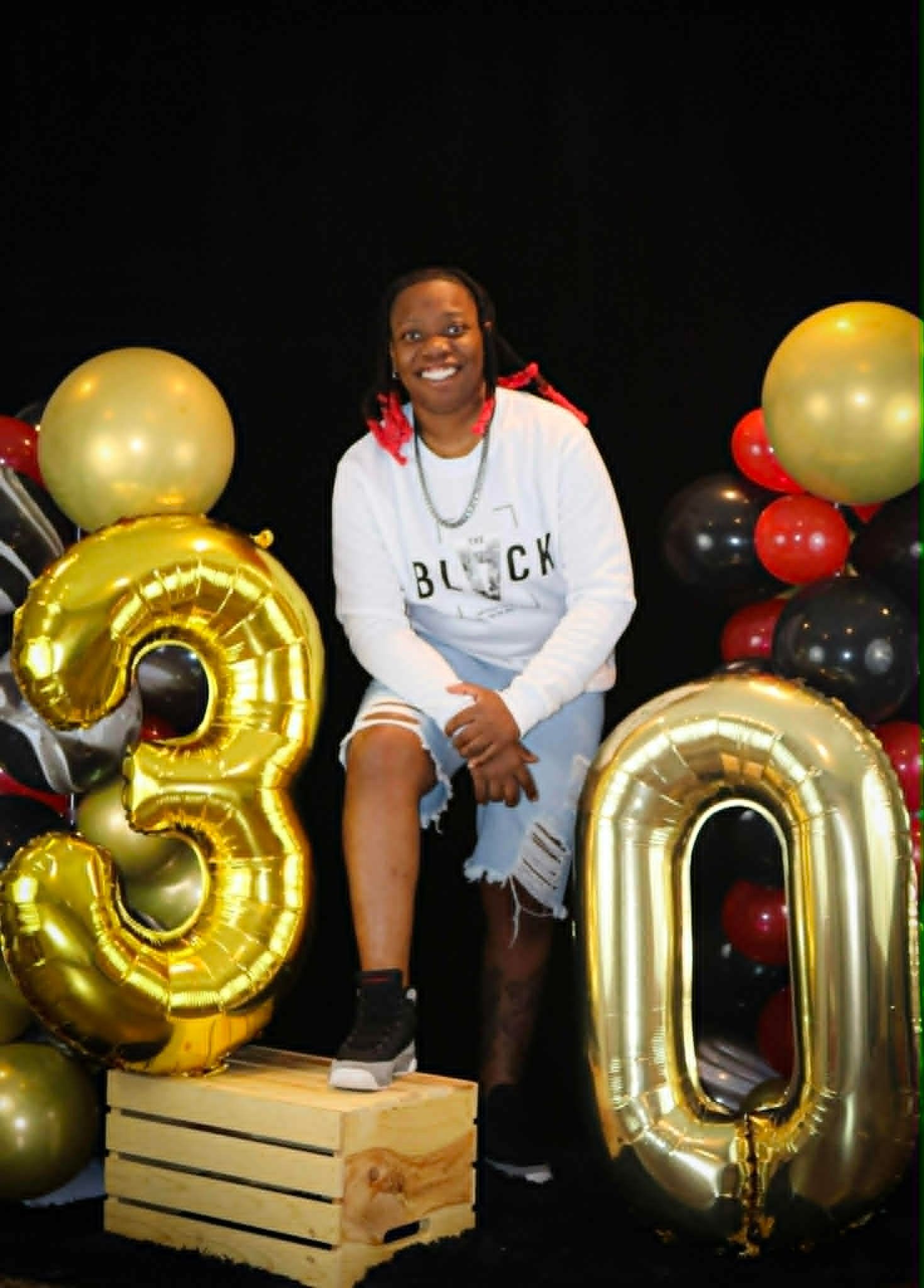 Person sitting on a crate between gold “3” and “0” balloons, smiling against a black backdrop.