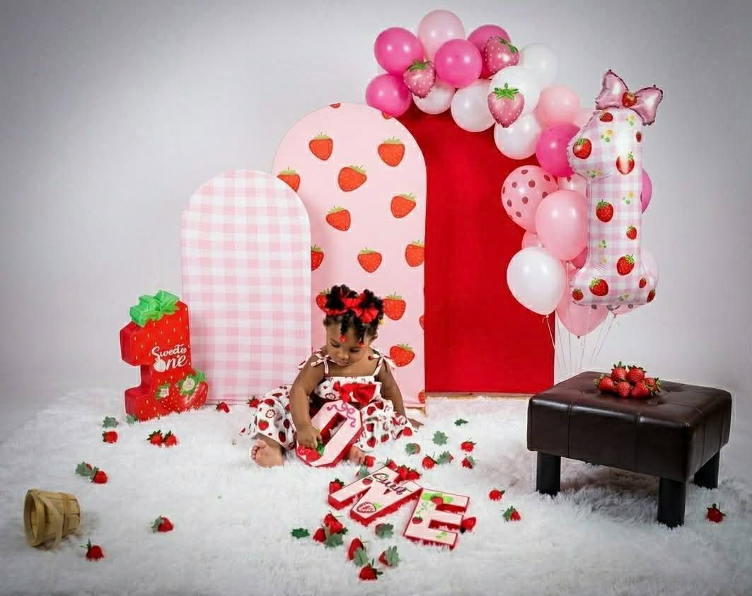 Festive baby in red outfit among balloons and heart decorations, surrounded by wrapped gifts and candy.