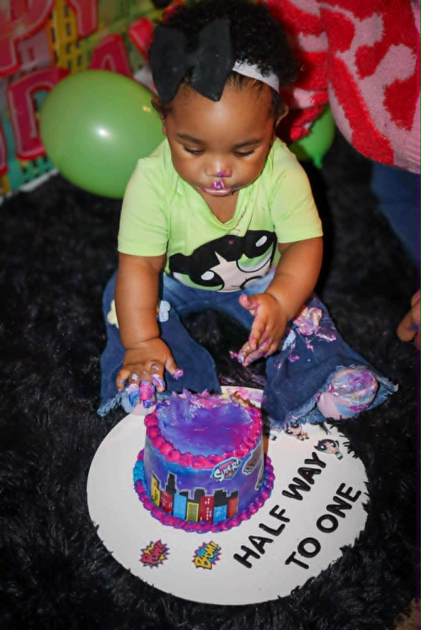 Child seated at a birthday cake on a white plate reading “HALF WAY TO ONE,” with balloons nearby.