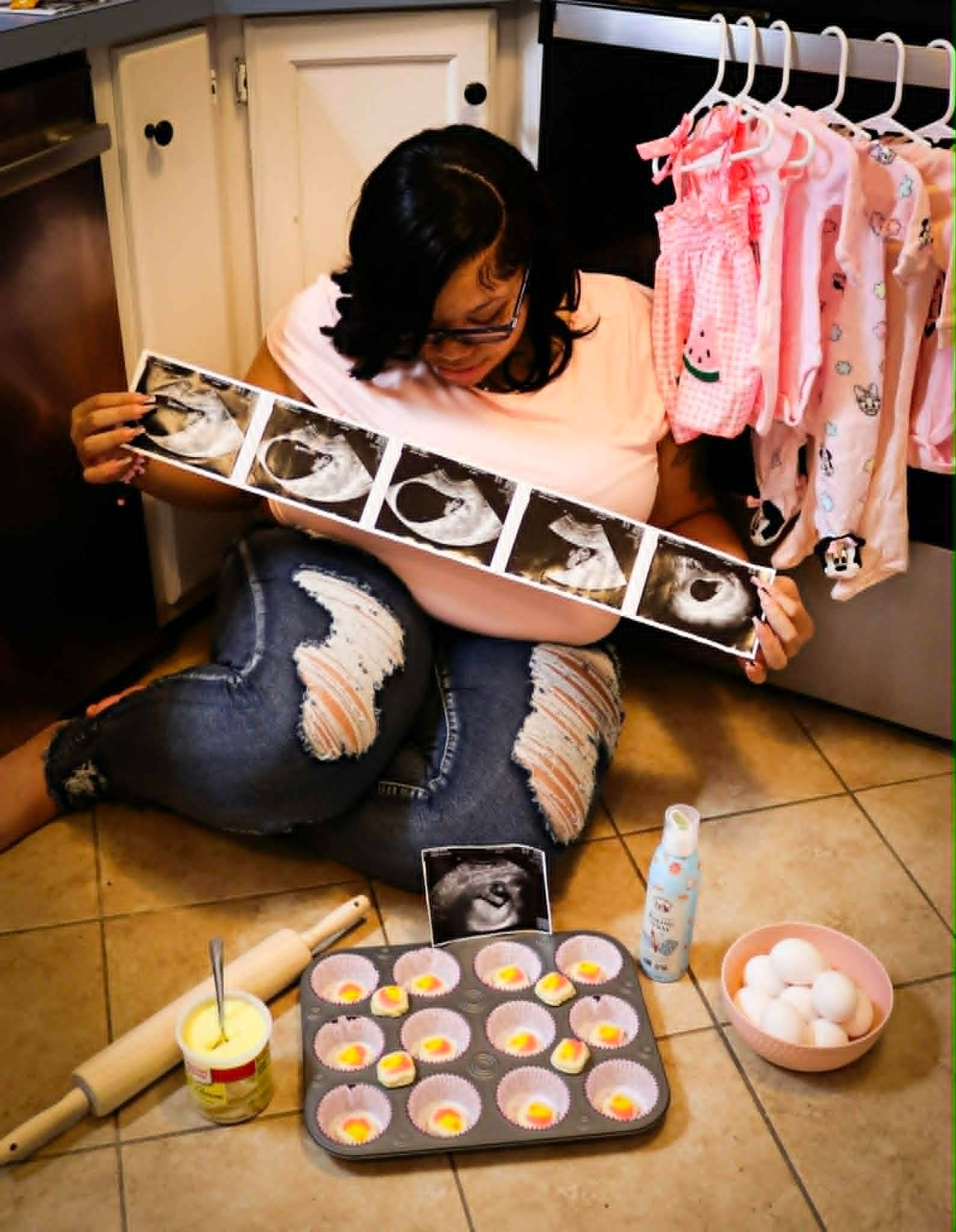 Person crouching on kitchen floor holding ultrasound printout, beside muffins, eggs, and baby clothes hanging nearby