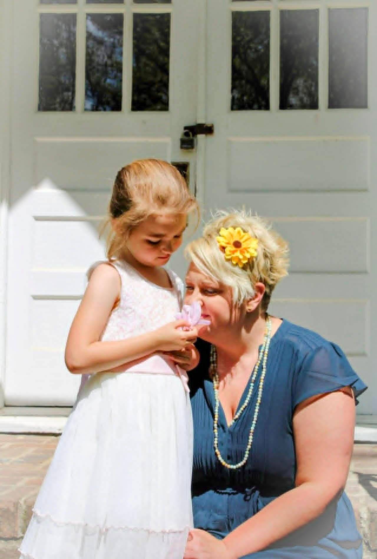 Young girl in white dress handing a flower to a seated woman on a front porch