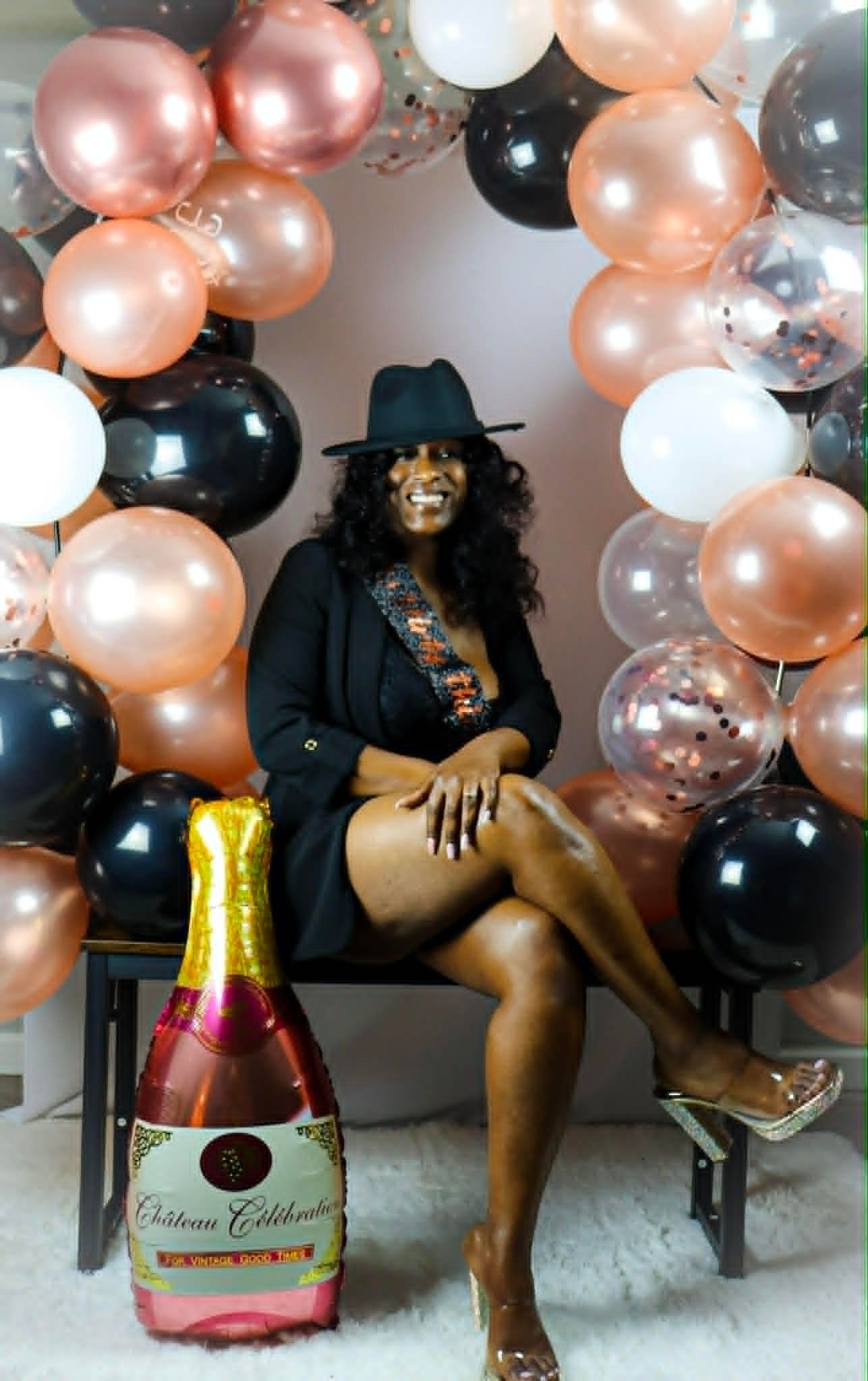 Person seated on a chair beneath a black, rose-gold, and white balloon arch, with a large champagne bottle in front