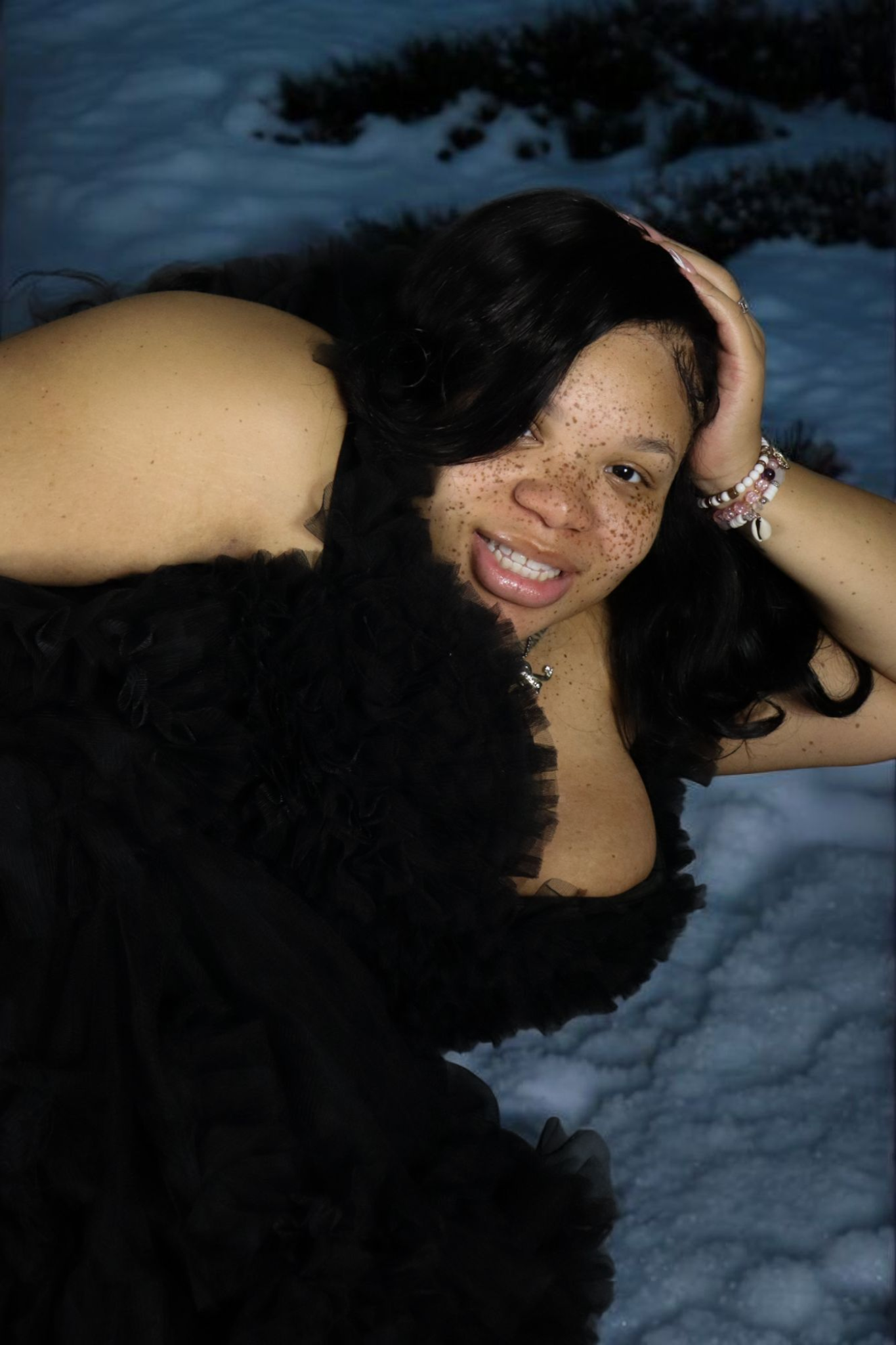 Woman in a black outfit posing on snow at night, smiling with one hand in her hair.