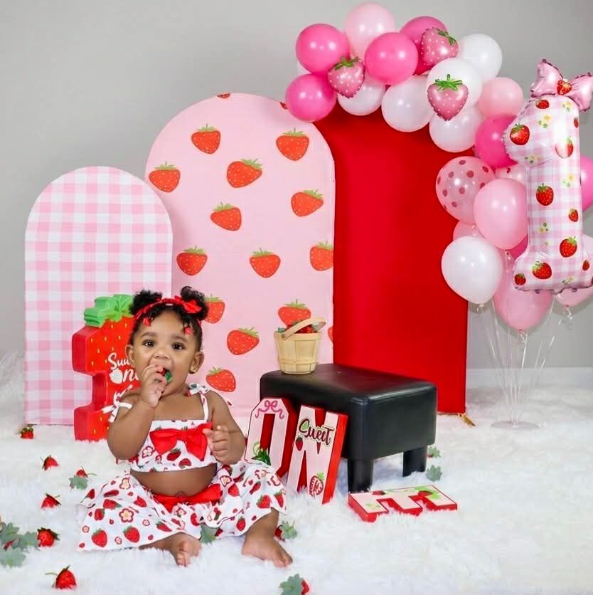 Child in red strawberry outfit sitting on a white rug at a pink strawberry-themed birthday party backdrop.
