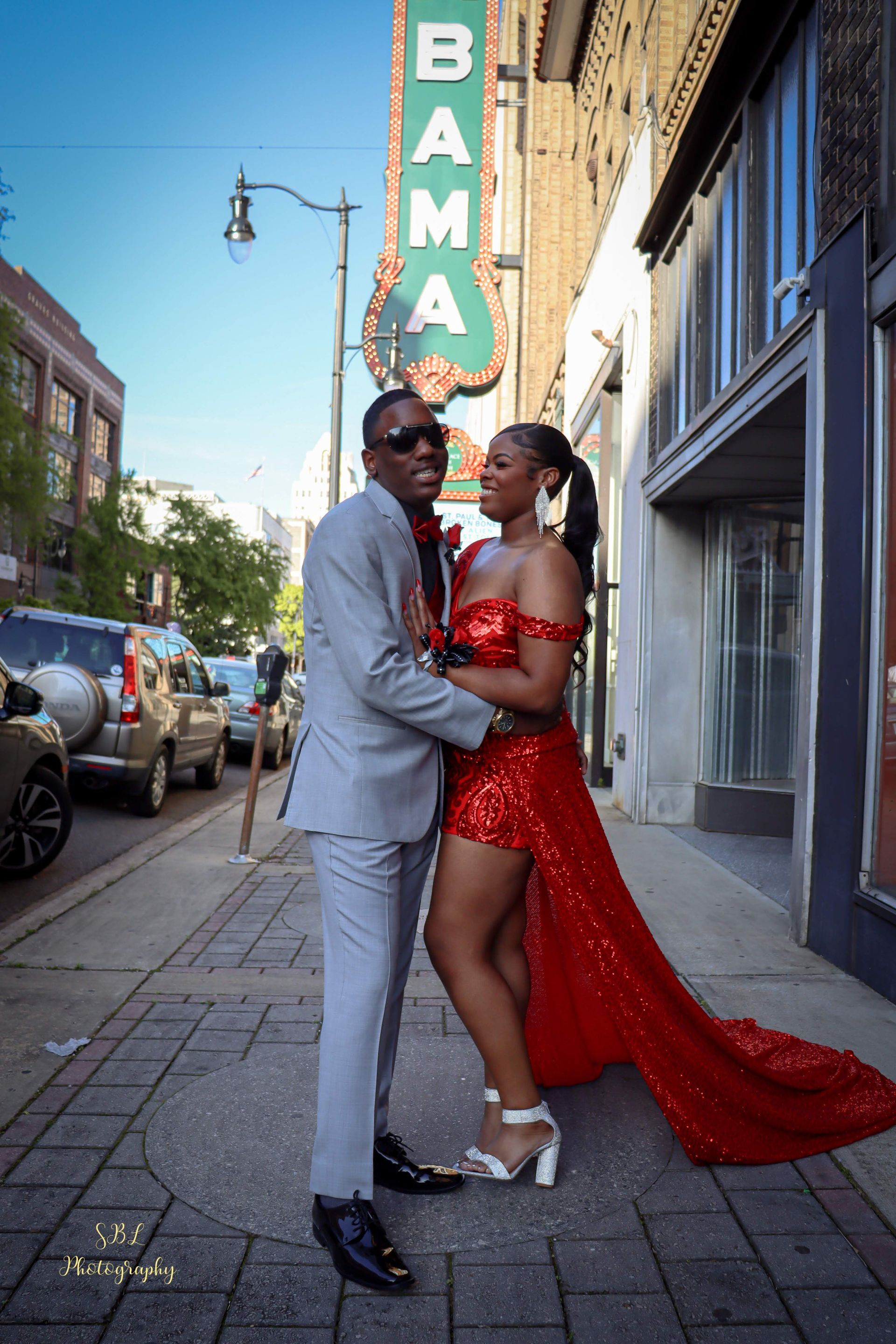 Couple posing on a city sidewalk, woman in red sequined dress with long train, man in light gray suit.