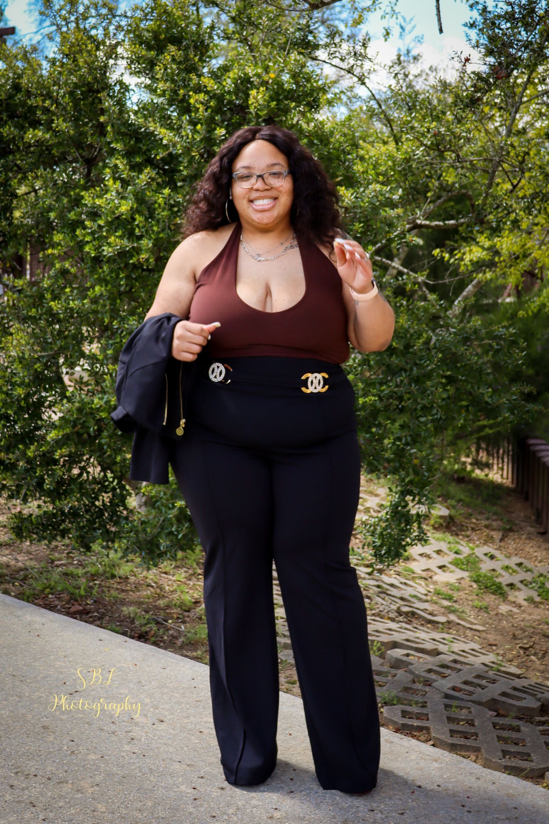 Woman smiling outdoors on a path, wearing a maroon top and black pants, holding a jacket.