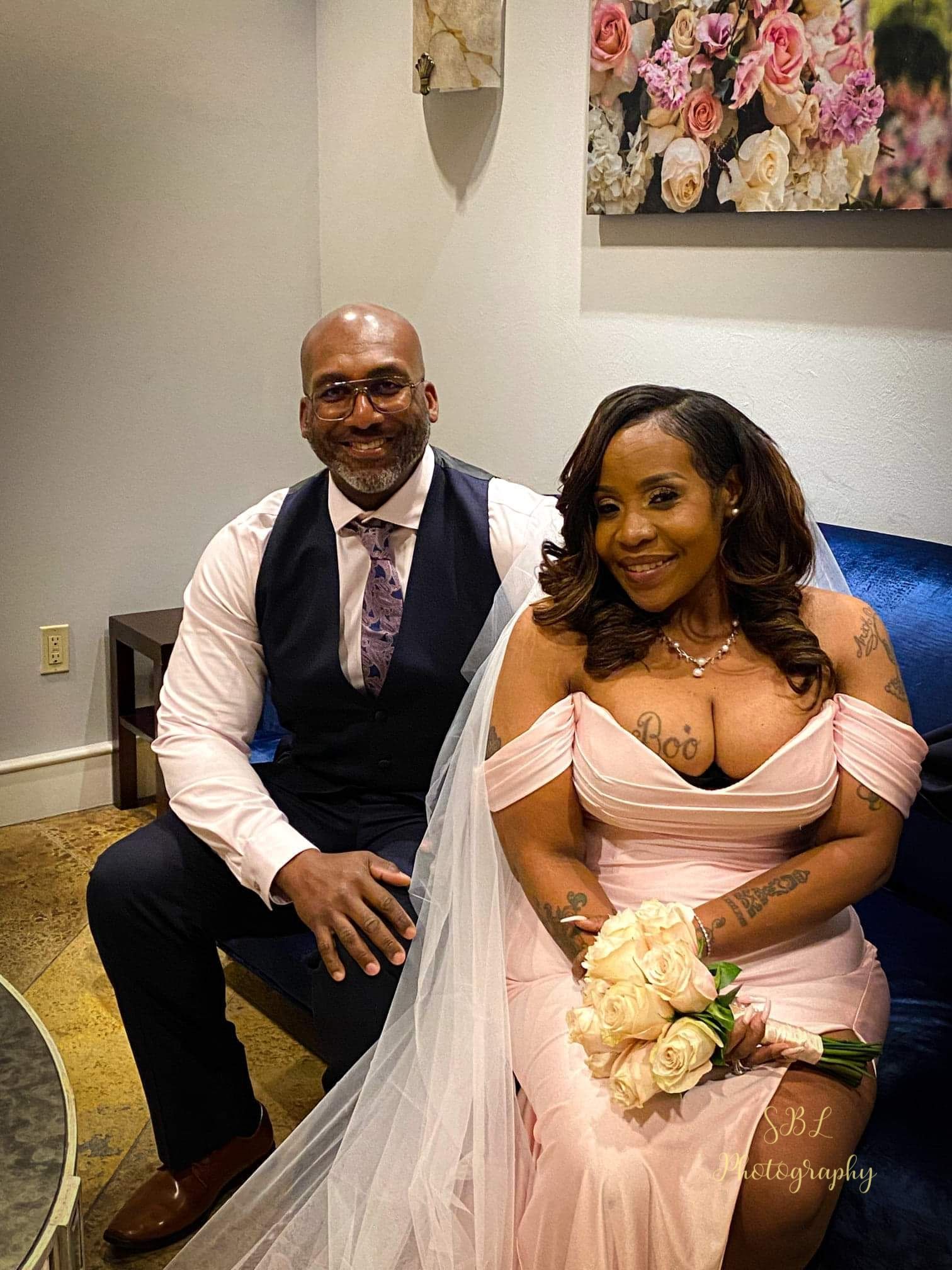 Two people in formal attire seated in a reception room, smiling, with a bouquet and wedding veil.