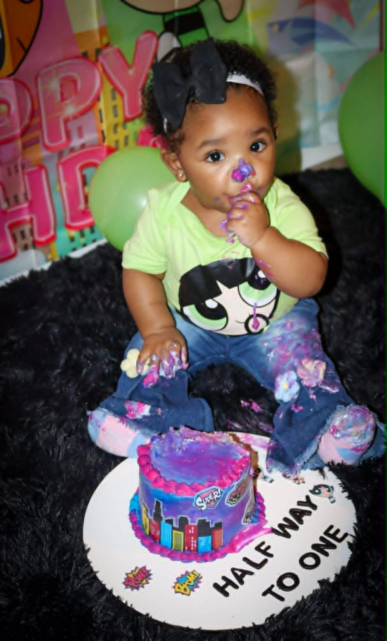 Child sitting with a cake on a birthday high chair, wearing a green shirt and purple pants.