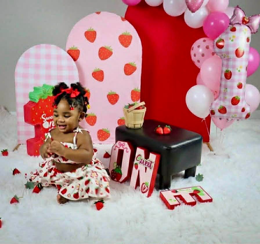 Toddler seated in a strawberry-themed birthday setup with pink balloons, gifts, and a cake table