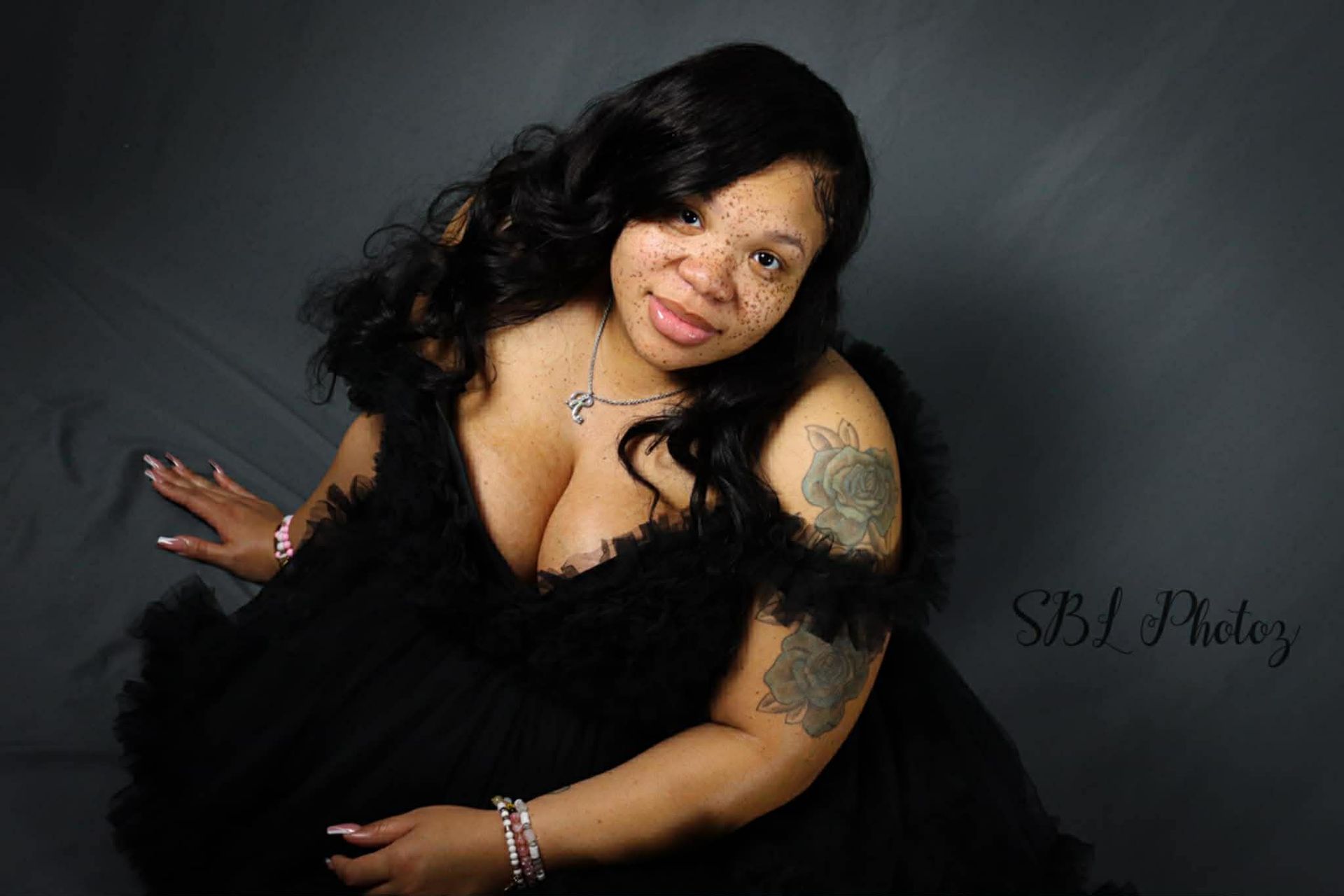 Woman in a black dress posing against a gray backdrop, smiling with curly dark hair and tattooed arm.
