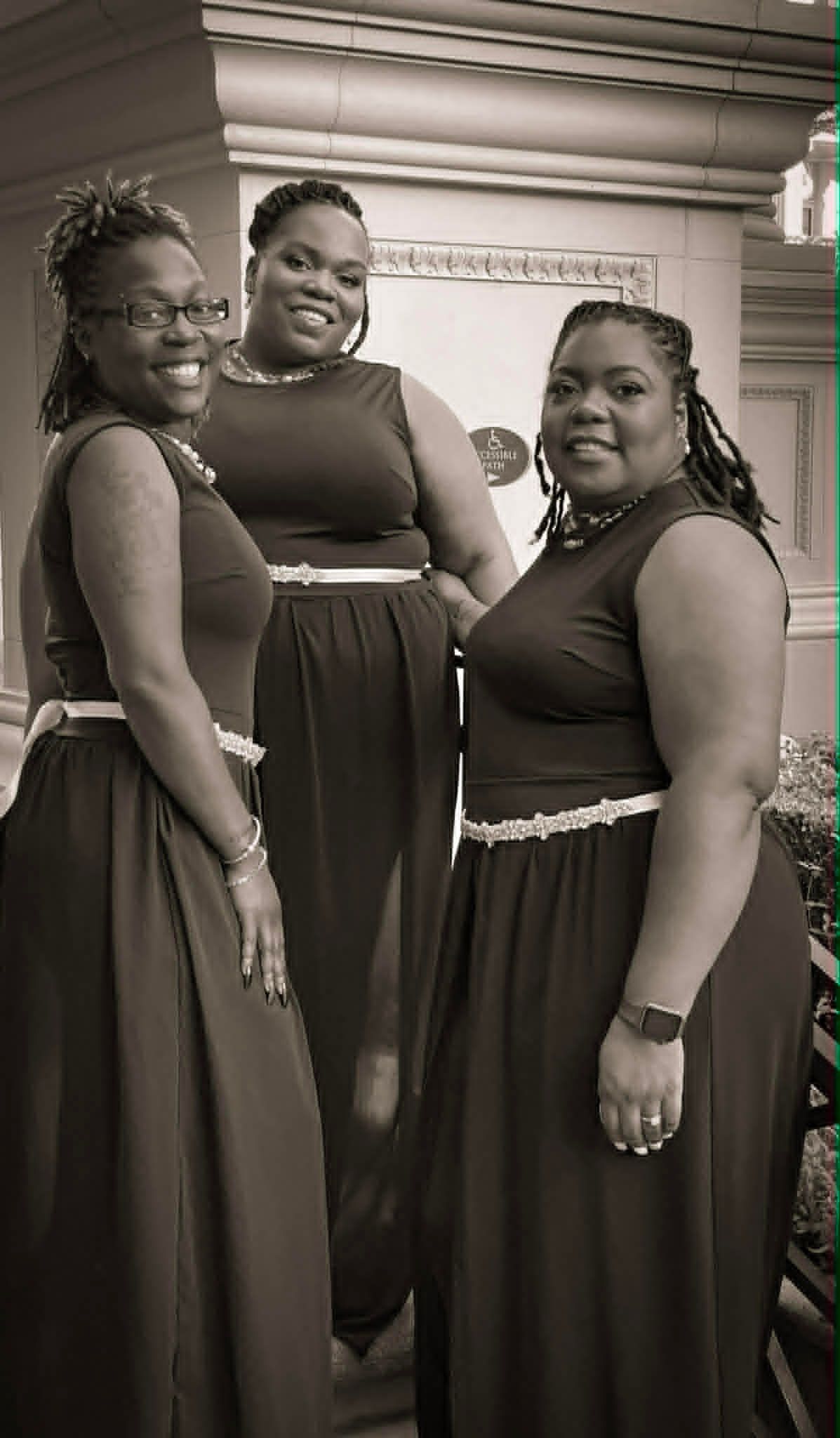 Three women in long dresses pose indoors, smiling at the camera.