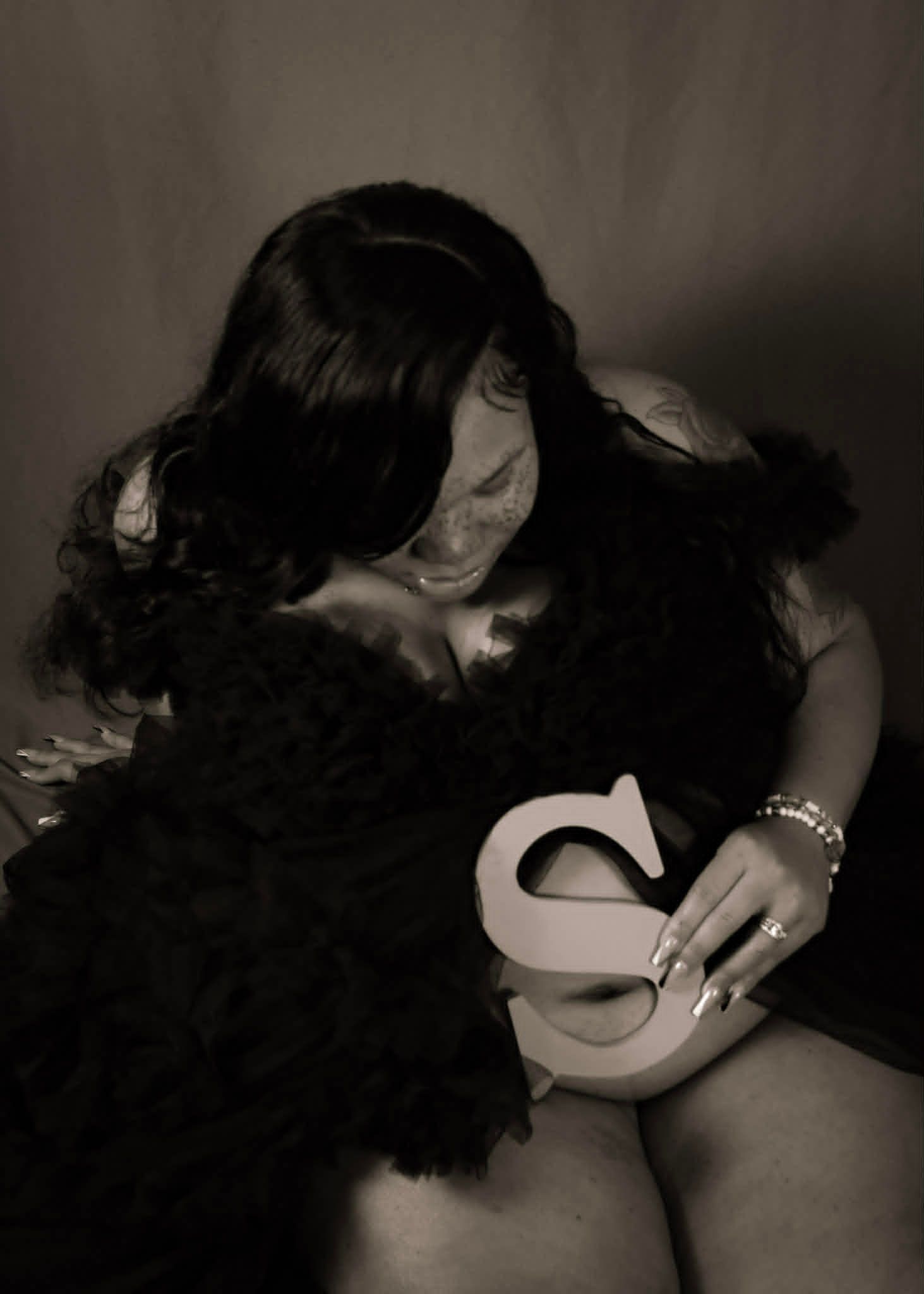 Woman with dark hair seated against a wall, holding a large white number 2 and wearing a black feathered wrap.