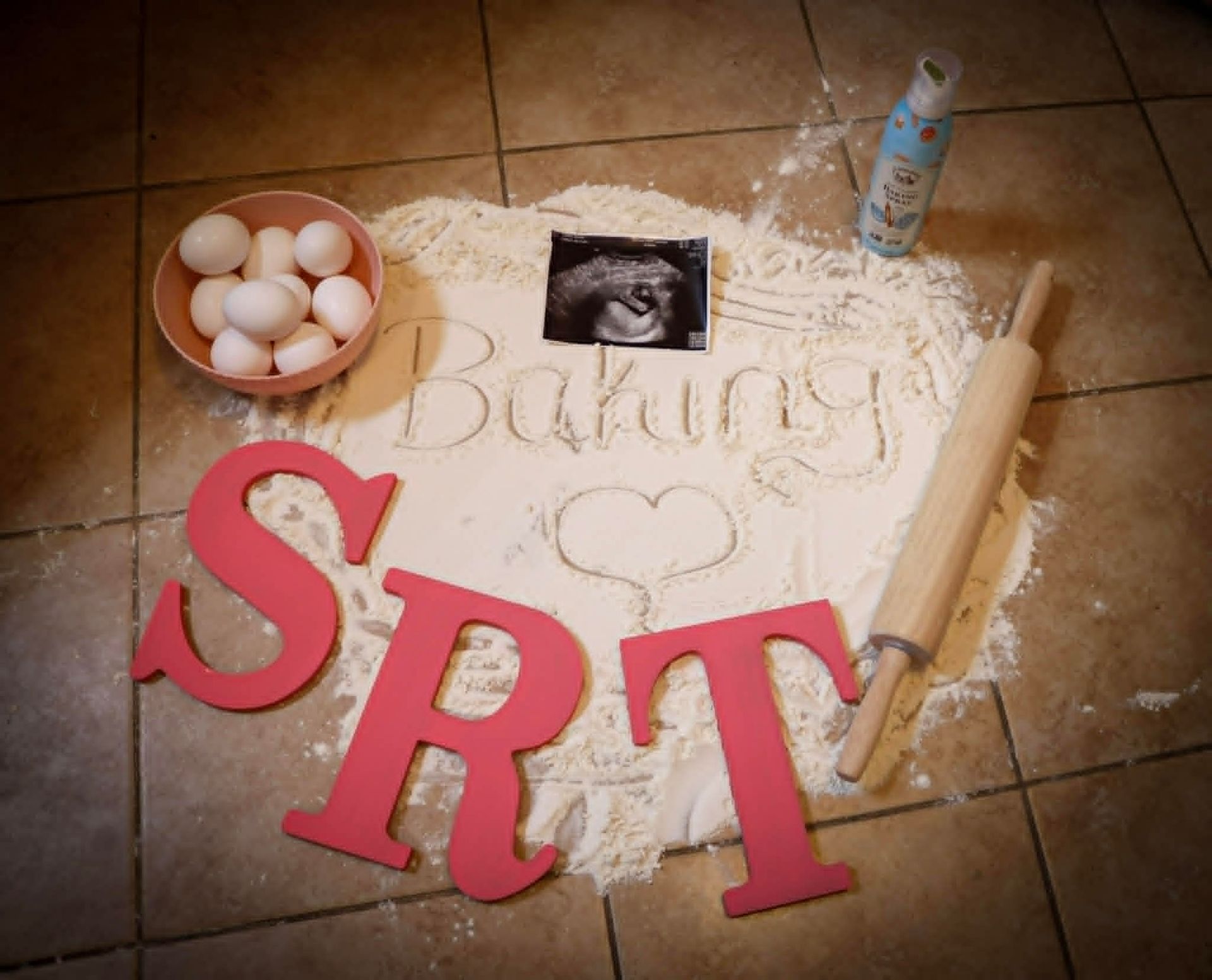 Baking-themed flat lay with eggs, flour, rolling pin, bottle, and red “SRT” letters on tile floor