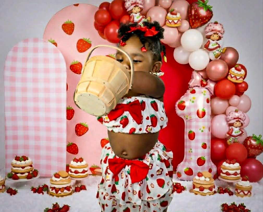 Child in a strawberry-themed outfit holding a milkshake, surrounded by pink balloons, cakes, and dessert decor.