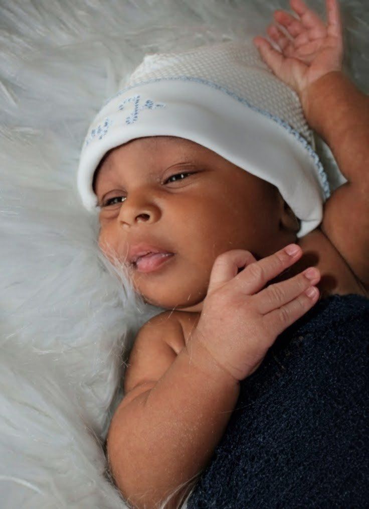 Sleeping baby in a white knit hat, lying on a fluffy white blanket with one hand near the face