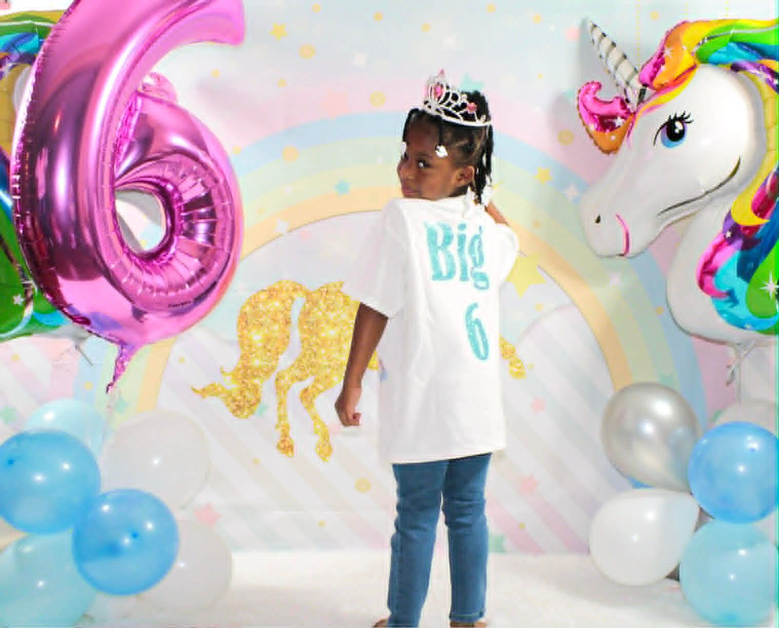 Child at a unicorn-themed birthday backdrop with balloons, wearing a tiara and “Big 6” shirt