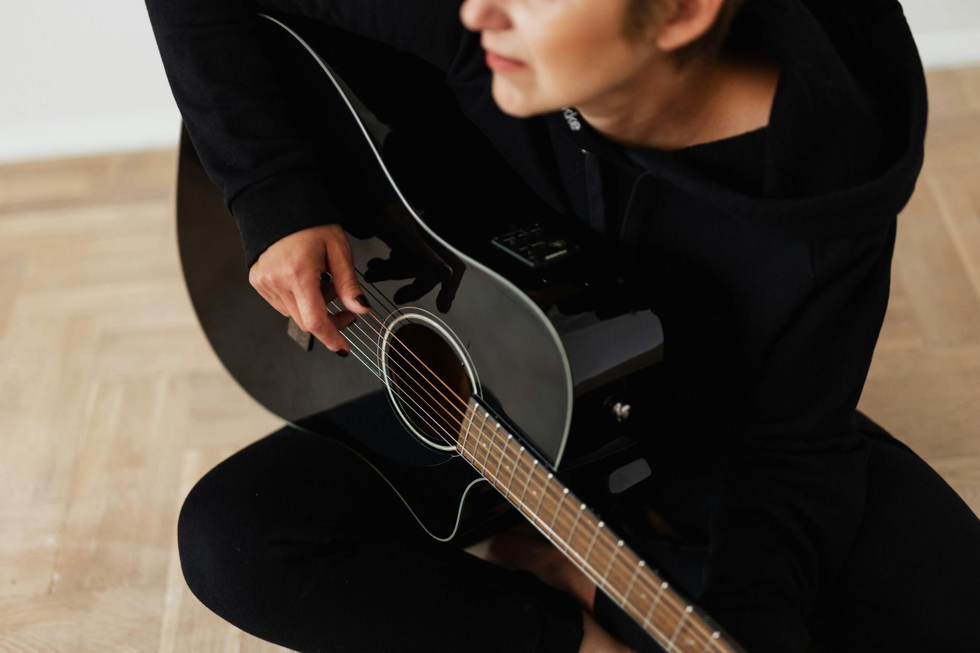 Een persoon in het zwart zit op een houten vloer en bespeelt een zwarte akoestische gitaar.