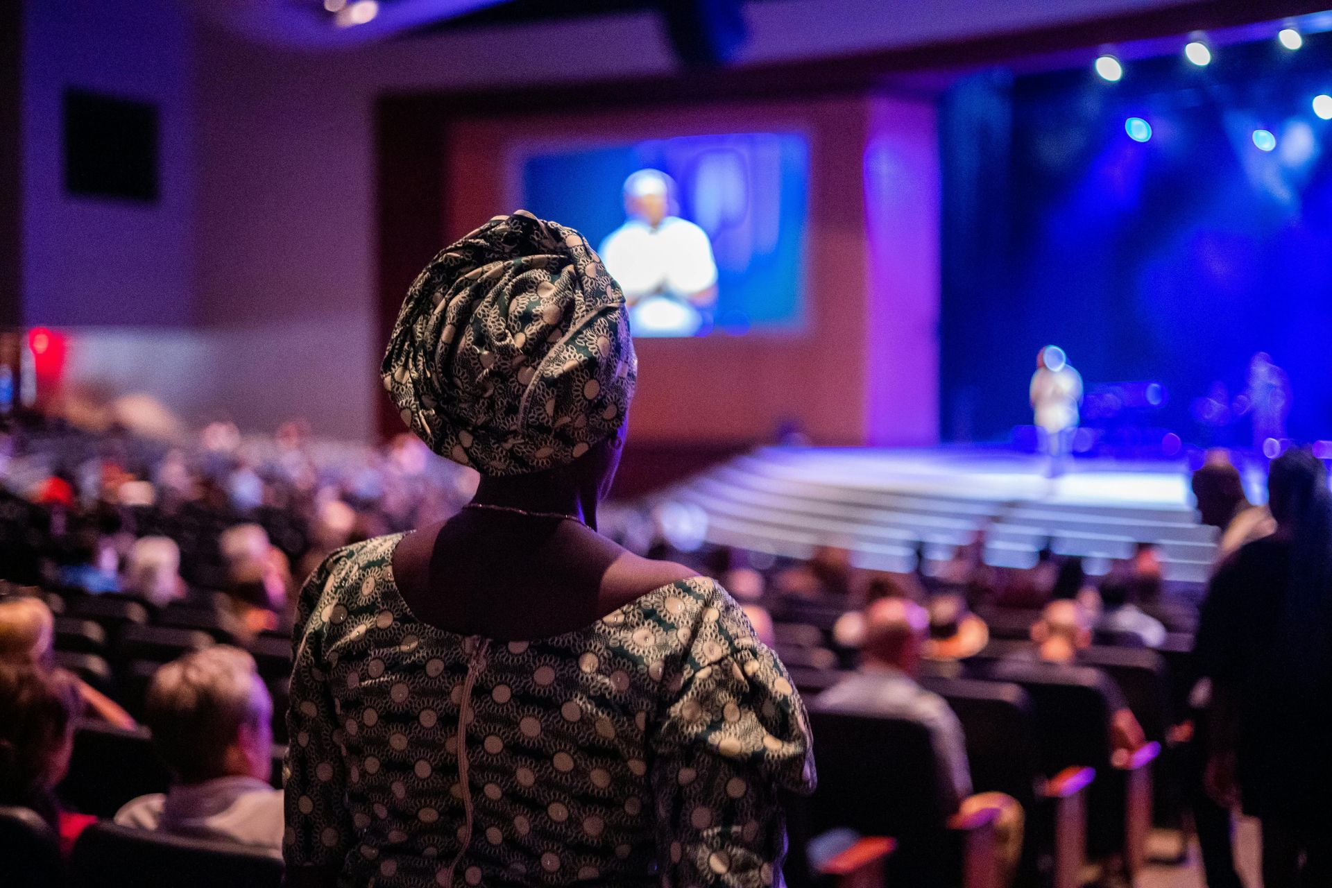 Een vrouw in Ankara met een hoofddoek op haar hoofd bekijkt een dienst op een podium met een groot scherm in een volle zaal.