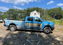 Blue GRIMBEE pickup truck parked on a dirt path near a house and trees under a blue sky
