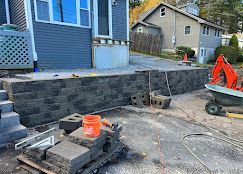 Backyard construction site with a newly built dark stone retaining wall, tools, and an orange excavator bucket