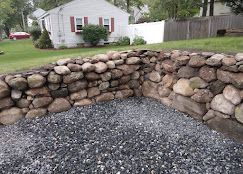 Stacked stone retaining wall around a gravel area in a yard, with a house and trees in the background