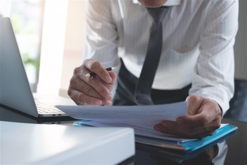 Man in dress shirt and tie, reviewing documents with a pen near a laptop and binder.