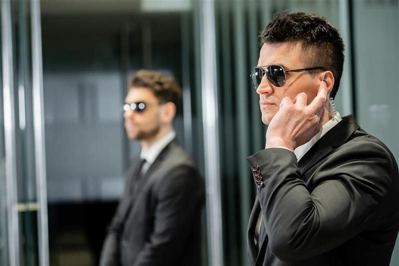 Man in a suit and sunglasses with an earpiece, standing in front of a mirror.
