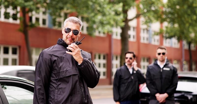 Man with walkie-talkie and bodyguards in front of a building, all wearing sunglasses and black jackets.
