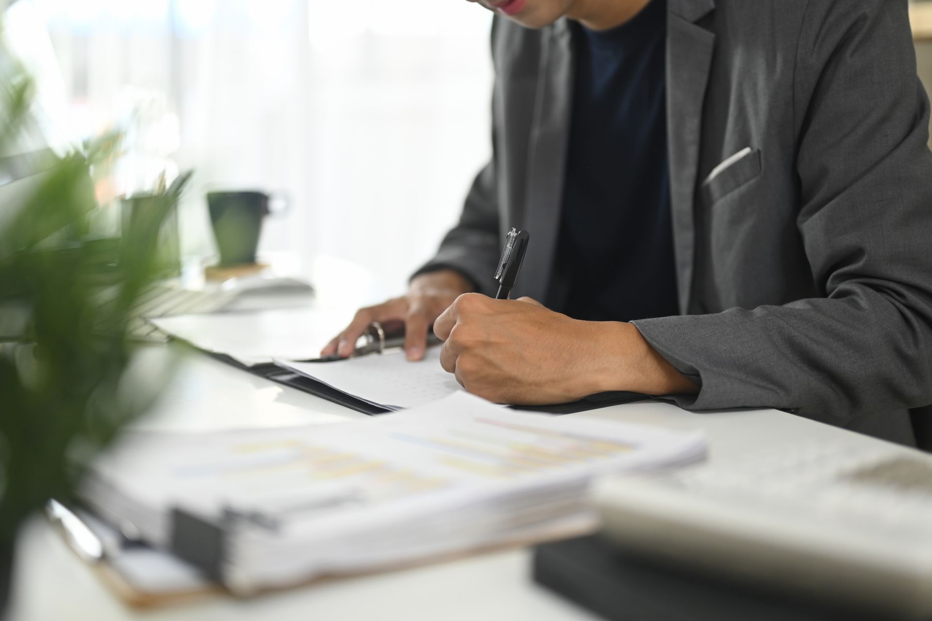 Person in a blazer writing on a paper at a desk. Papers and a plant are nearby.